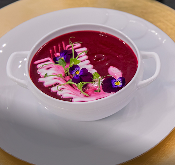 Krem z buraka w białej miseczce na talerzu, udekorowany śmietaną, ziołami i jadalnymi kwiatami