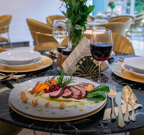 Elegancko podane danie z plastrami mięsa i warzywami na białym talerzu, obok kieliszek czerwonego wina i złote sztućce