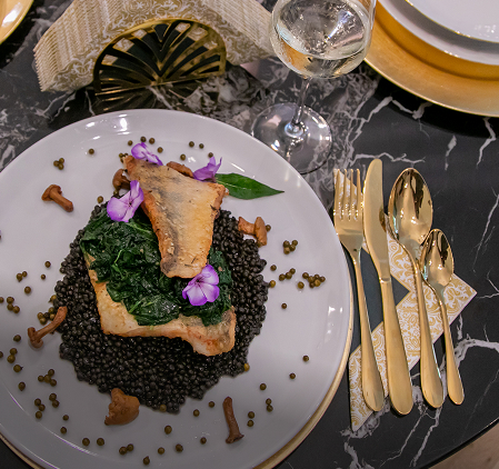 Elegancko podane danie z rybą na czarnej soczewicy ze szpinakiem i jadalnymi kwiatami, obok złote sztućce